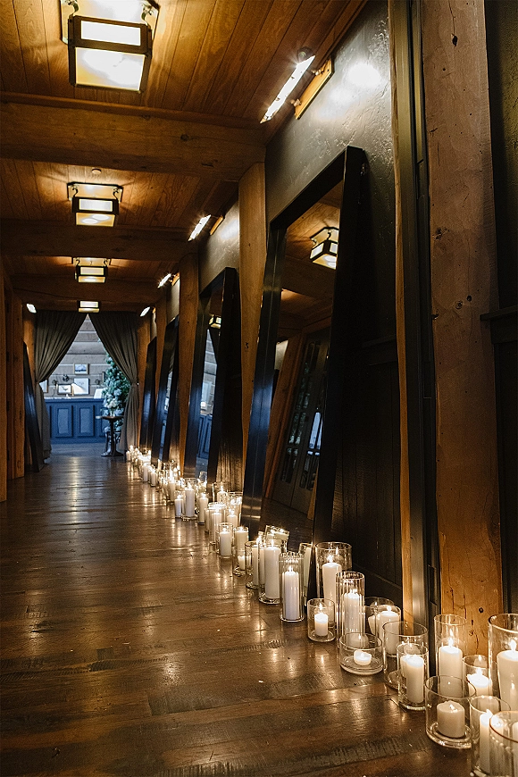Candlelit wedding aisle lined with wedding aisle candles in glass cylinder vases and floor mirrors, set in a rustic wood hallway with draped curtains