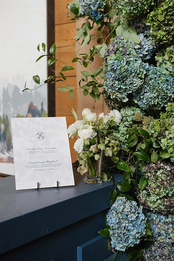 Wedding bar menu with signature cocktail menu sign on a stand beside hydrangeas and greenery garland at a wooden bar counter