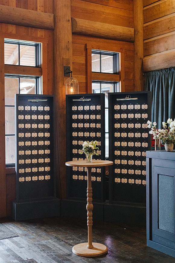 Wedding seating chart with escort card display boards and clip-on lights on a bistro table, framed by wood-paneled walls and windows.