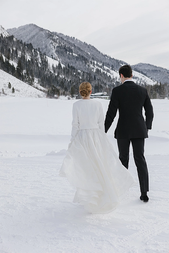 Couple portrait of bride and groom walking away in snow, her long-sleeve full skirt dress flowing beside his tuxedo, with mountains and pines under an overcast sky
