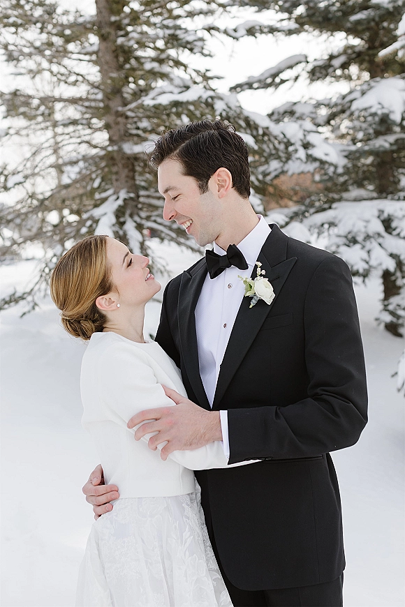 Couple portrait of a winter wedding couple embracing in falling snow, groom in black tuxedo with white rose boutonniere by evergreens