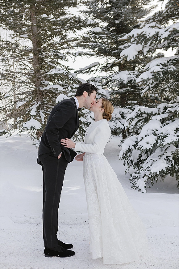 Wedding kiss portrait of bride in long sleeve dress and groom in black tuxedo sharing a kiss in a snowy evergreen forest backdrop