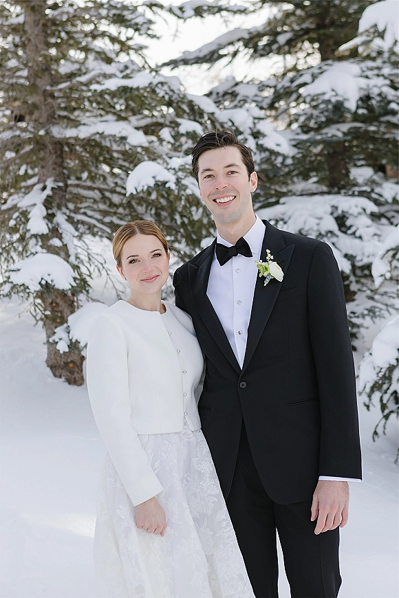 Couple portrait of bride in a white coat and groom in black tuxedo with bow tie, standing in a snowy evergreen forest