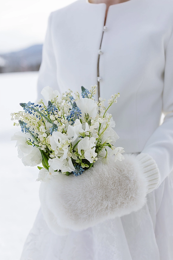 Bridal bouquet of white and blue flowers with lily of the valley, held with white gloves and fur muff against snowy mountains