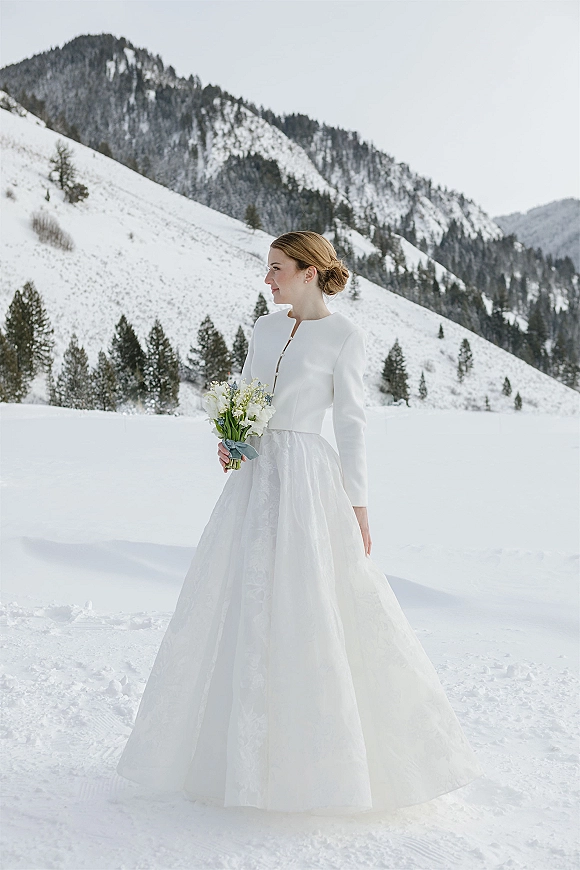Bridal portrait of a winter bride holding a white and green bouquet, side profile in a long sleeve jacket with snowy mountains and pines behind