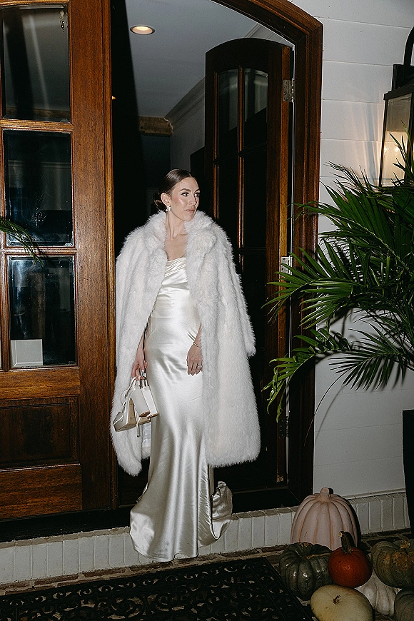 Bridal portrait of a bride in a satin slip wedding dress with faux fur coat, holding heels and a handbag by a porch wood door.