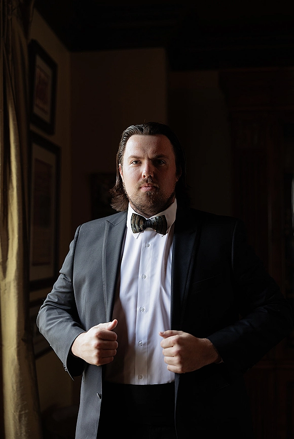 Groom portrait in a tuxedo, adjusting his suit jacket and bow tie in window light beside curtains and framed wall art indoors