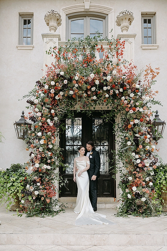 Couple portrait of bride in off the shoulder wedding dress and groom in black tuxedo beneath a rose floral arch at a villa doorway