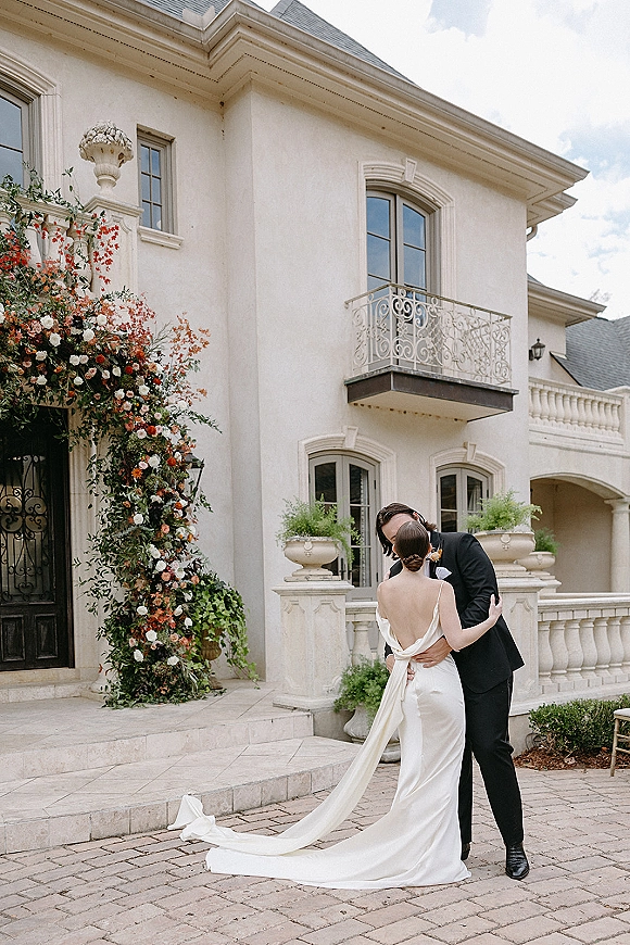 Wedding kiss portrait of bride and groom kissing beneath a rose greenery arch on stone steps outside a stucco villa with French doors