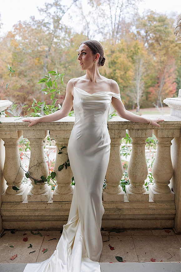 Bridal portrait of a bride in an off the shoulder wedding dress with a draped neckline, posing by a stone balustrade on an ivy terrace in autumn