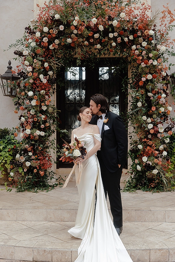 Couple portrait of groom kissing bride’s forehead as she holds a rose bouquet beneath a floral arch at a wrought iron doorway