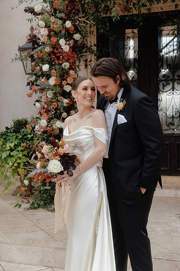 Couple portrait of bride and groom posing under an asymmetrical floral arch, bouquet in hand, by stucco wall and wrought iron doors