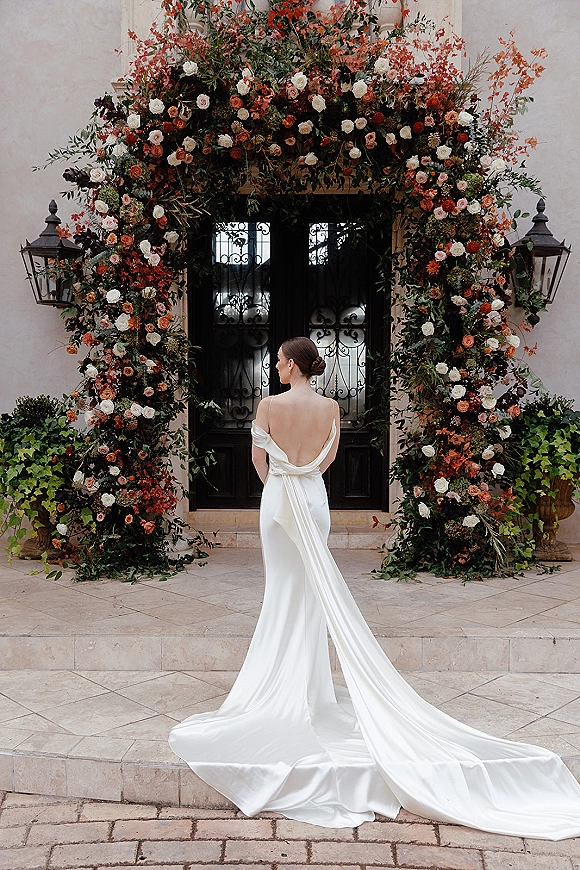 Bridal portrait of a bride from behind in a backless wedding dress with long train beneath a rose floral arch at ornate double doors