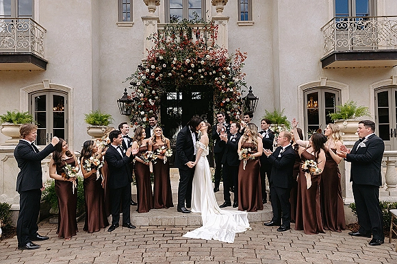 Wedding party portrait of bride and groom kissing as bridesmaids and groomsmen cheer on mansion steps beneath a floral arch and greenery garland