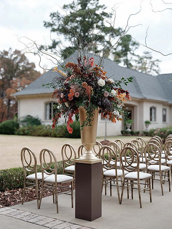 Ceremony aisle decor with aisle floral arrangement in a gold urn, fall foliage, greenery and curly willow beside courtyard chairs and trees.
