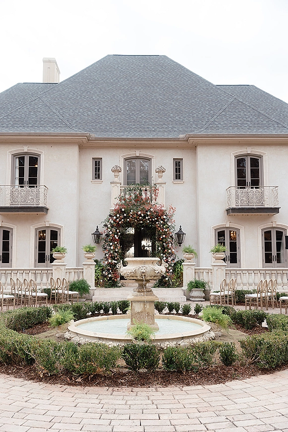 Ceremony setup with outdoor ceremony seating facing a stone fountain, framed by a floral arch and white estate courtyard facade with lanterns