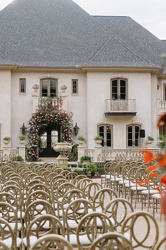 Ceremony setup for an outdoor wedding ceremony with gold round-back chairs facing a floral arch in an estate courtyard by a stone fountain.