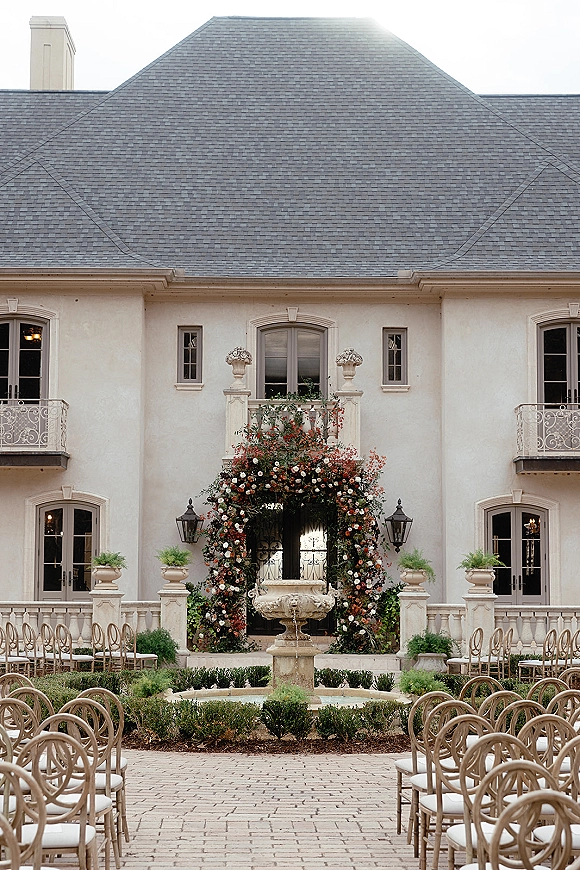 Ceremony setup for an outdoor wedding ceremony with a floral arch by a stone fountain, wooden chairs in a manor courtyard setting