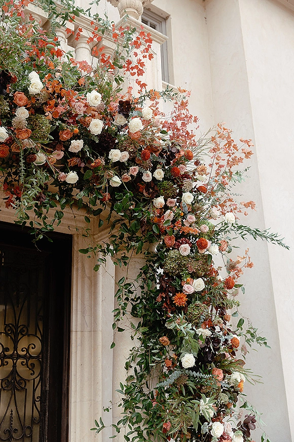 Wedding floral installation of roses and greenery framing a stone doorway with vines cascading over a wrought iron gate at a villa entrance