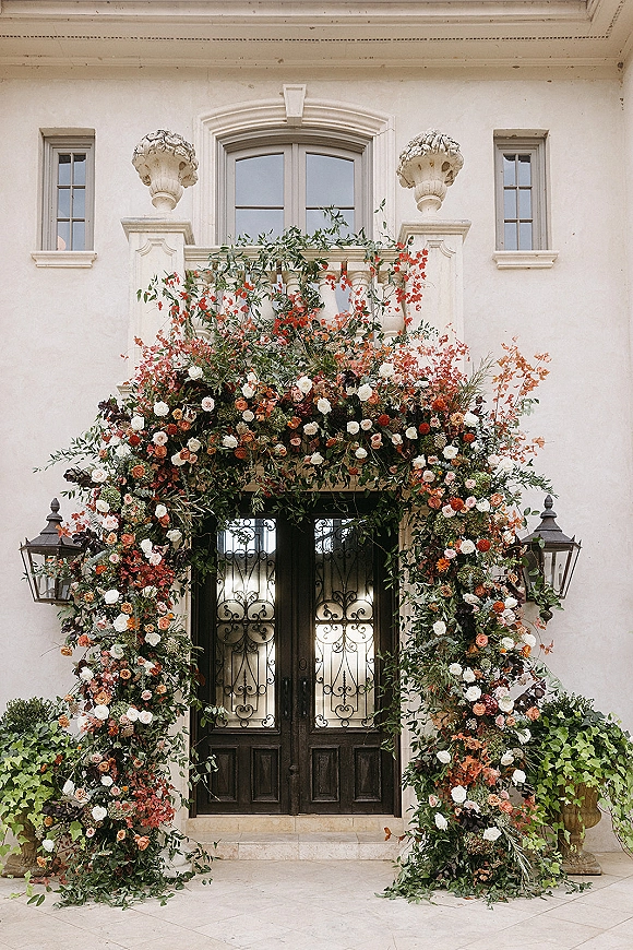 Wedding floral arch framing iron front doors with roses and greenery, climbing vines and black lantern accent at a stucco villa entryway