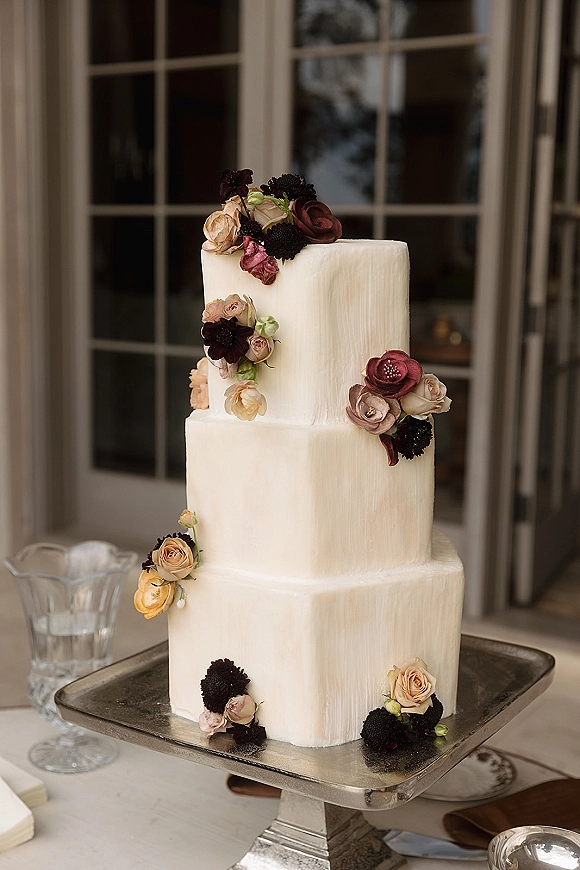 Wedding cake with a square wedding cake design in white buttercream, topped with cascading sugar roses on a metal stand by window panes
