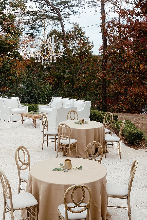 Outdoor lounge decor with white sofas and wedding lounge seating around round tables under chandelier and string lights on a stone patio
