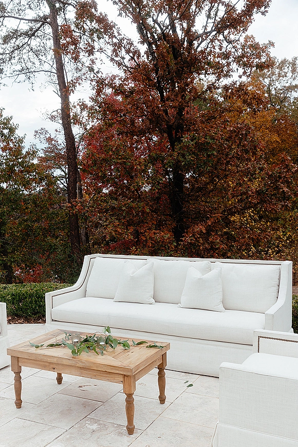 Wedding lounge seating with a white sofa and wooden coffee table topped with greenery garland on a stone patio under autumn trees