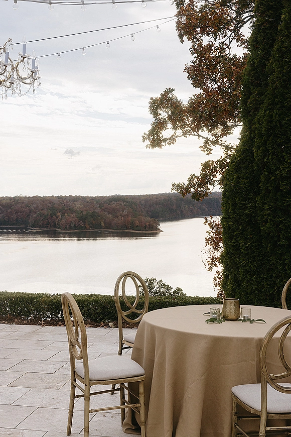 Wedding reception patio with a round table in taupe linen, gold chairs, greenery garland, and glass votives under string lights by the lake