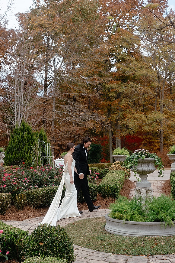 Couple portrait of bride and groom walking hand in hand on a manicured garden path, her long dress train trailing beside pink flowers