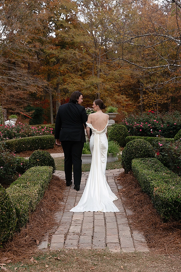 Couple portrait of bride and groom walking away holding hands, wedding dress train trailing on brick walkway in formal garden with fountain