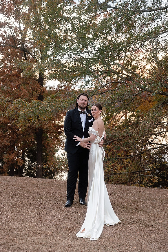 Couple portrait of bride and groom embracing, her off-shoulder wedding dress and his tuxedo, by a lakeside lawn at sunset amid autumn trees