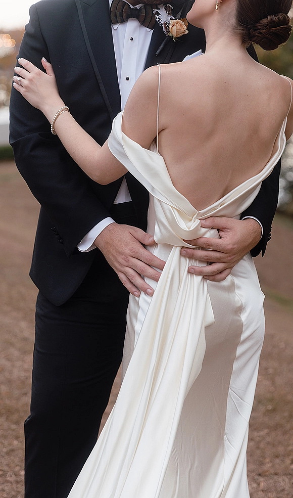 Couple portrait of bride and groom embrace, her off-shoulder satin low-back gown and his tuxedo with rose boutonniere on a walkway with bokeh lights