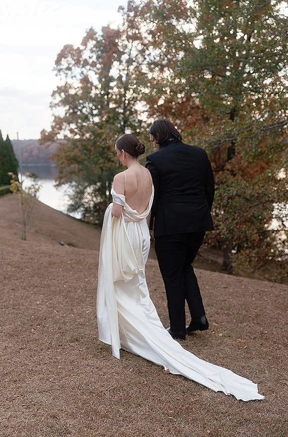 Couple portrait of bride and groom walking away, her backless satin dress train flowing on a lakeside lawn with autumn trees behind