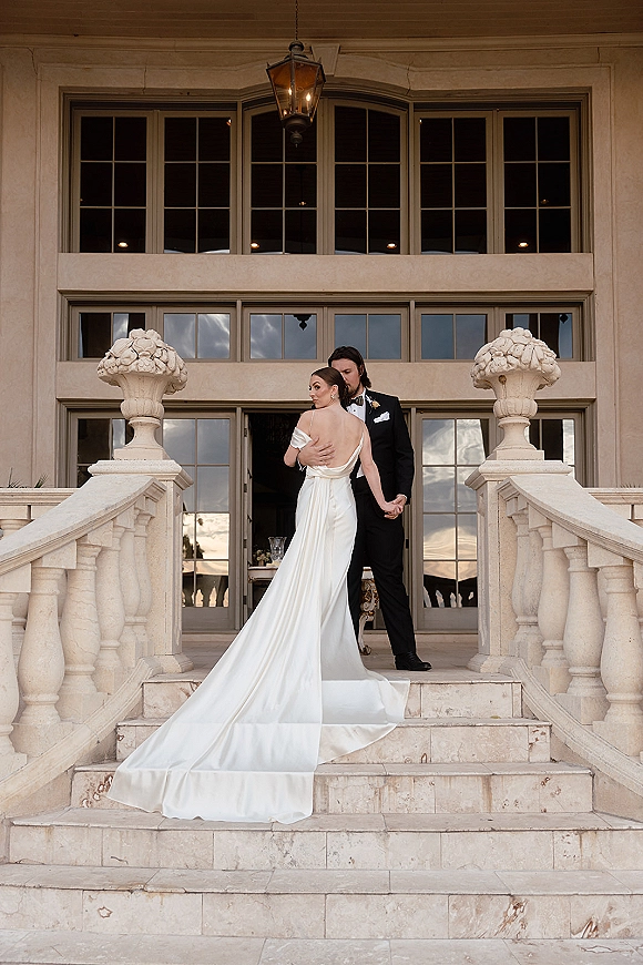 Couple portrait of bride and groom on steps, holding hands as her long satin train trails on a stone staircase with lanterns behind