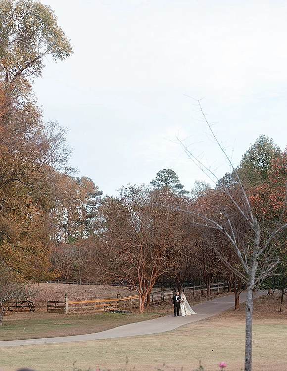 Couple portrait of bride and groom walking hand in hand, bride with veil and bouquet, groom in tuxedo on a tree-lined path in fall foliage