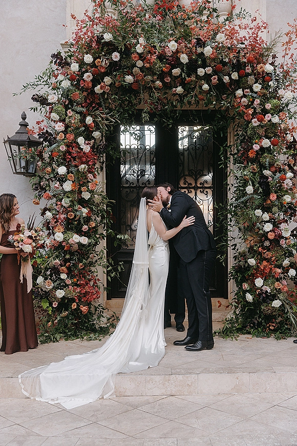 Wedding kiss beneath a rose and greenery floral arch at a stone doorway, bride’s veil and train flowing as bridesmaid watches nearby