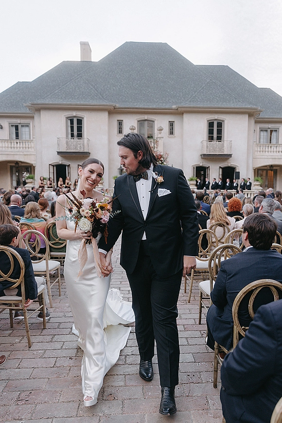 Wedding recessional as bride and groom walk the aisle holding hands, bride with bouquet, guests on stone pavers by an estate building under overcast sky
