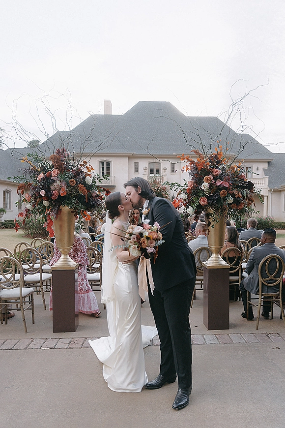 Wedding kiss as bride in satin dress and veil holds bouquet, groom in black tuxedo, in courtyard aisle with guests and urn florals