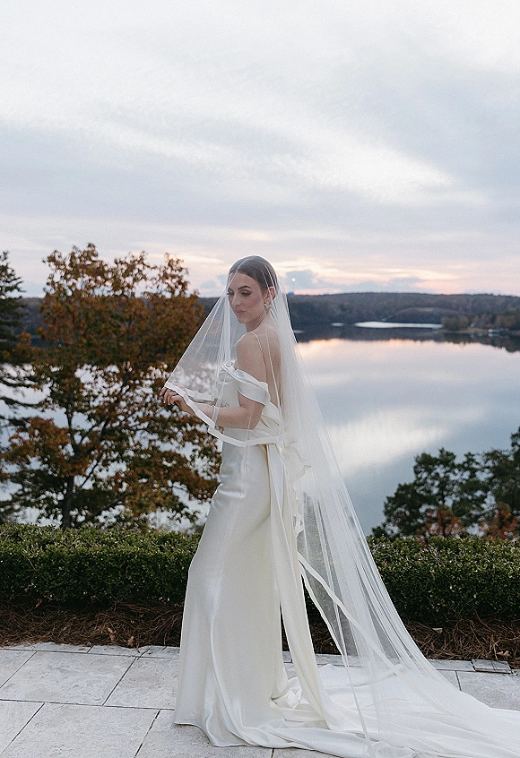 Bridal portrait of a bride in veil, cathedral veil draped over her face as she looks back on a stone terrace by a lake under clouds