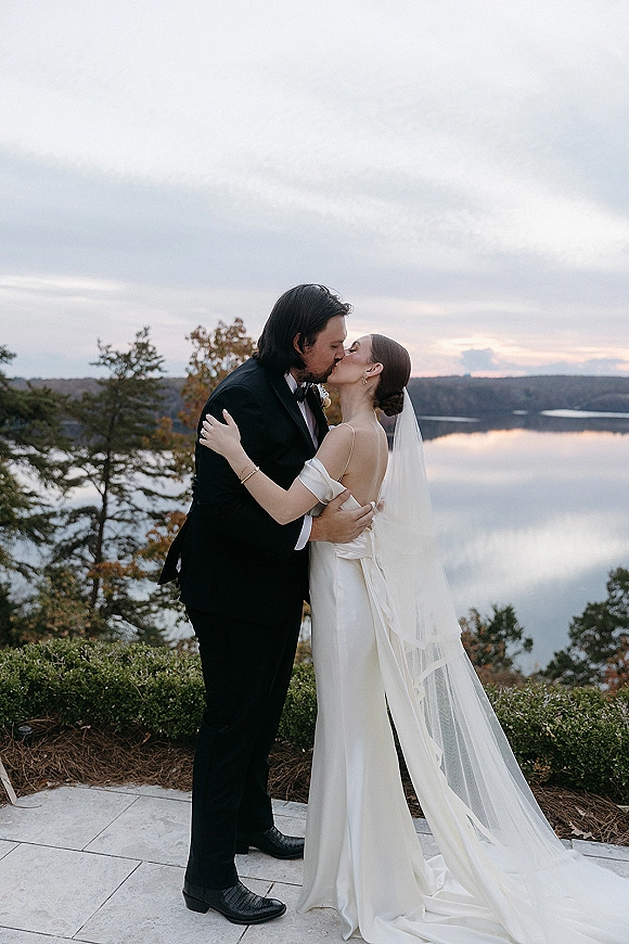 Wedding kiss portrait of bride and groom kissing, her long veil blowing slightly as they embrace on a stone patio by a lake