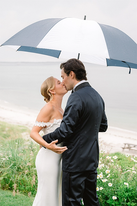 Wedding kiss portrait of bride and groom kissing under a white umbrella, her strapless lace dress by the ocean on an overcast beach