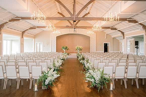 Ceremony setup with white florals and greenery aisle arrangements, candles and lanterns lining a wood-floor hall with exposed beams