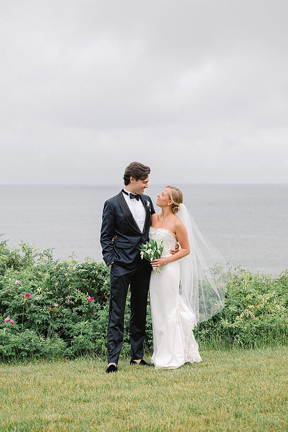 Couple portrait of bride and groom smiling, her veil blowing and bouquet in hand, in tuxedo beside ocean under overcast sky
