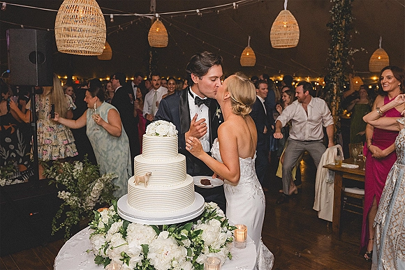 Cake cutting moment as bride in strapless gown and groom in black tuxedo kiss by a tiered white cake under string lights in a tent reception