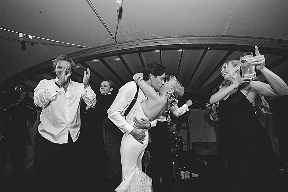 Wedding kiss moment as groom in suspenders dips bride in strapless lace dress on tent dance floor under string lights, guests cheering