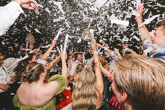 Wedding dance floor erupts in a wedding confetti blast as guests dance with hands up under wicker pendant lights at an outdoor night reception