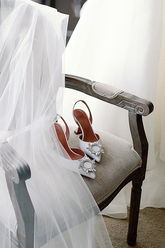 Bridal shoes in white slingback heels with crystal shoe clips, draped with a tulle veil on an upholstered wooden chair against a white wall
