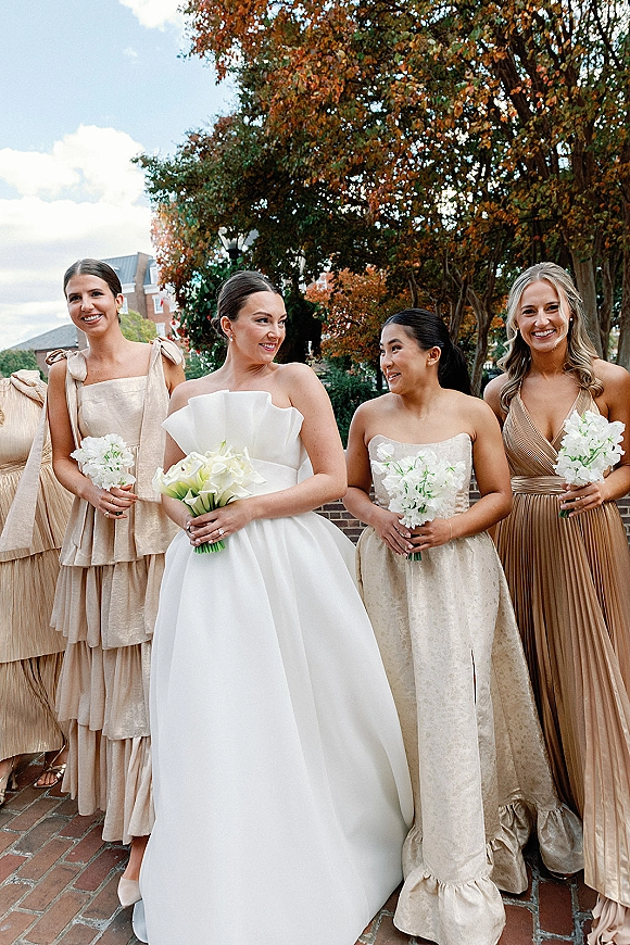 Bridesmaid group photo of bride with bridesmaids in champagne dresses holding white calla lily bouquets on a brick walkway with autumn trees