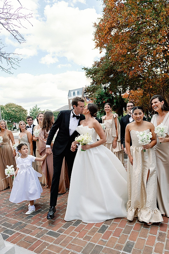 Wedding kiss as bride in strapless ball gown and groom in black tuxedo walk with bridal party on a brick street in autumn