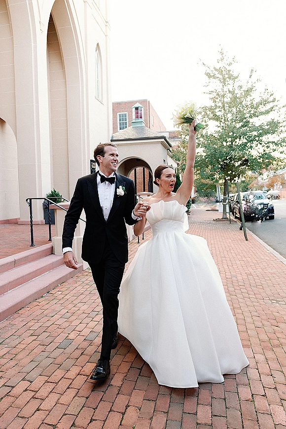 Newlywed couple in a wedding recessional, holding hands as the bride lifts her bouquet outside a church on a brick sidewalk